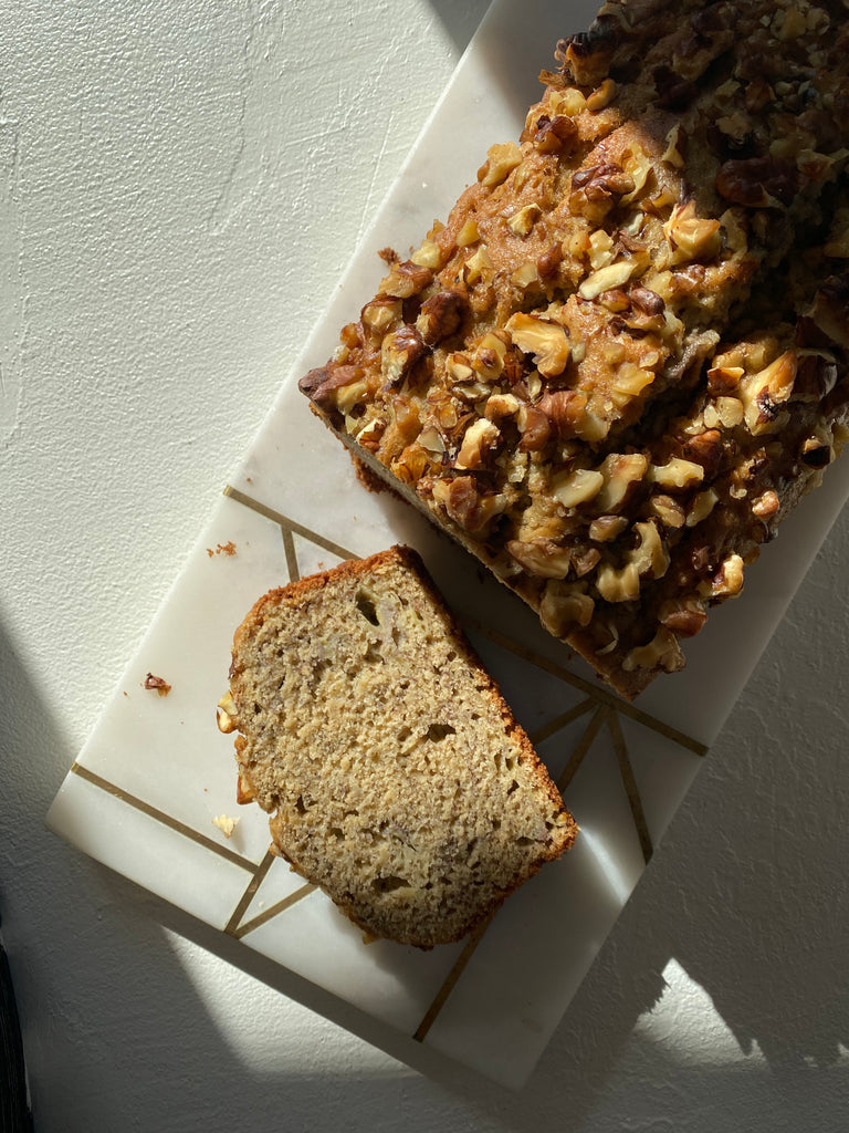 Top view of banana bread loaf with nuts on top, with a slice cut off lying in front of it on a white chopping board.