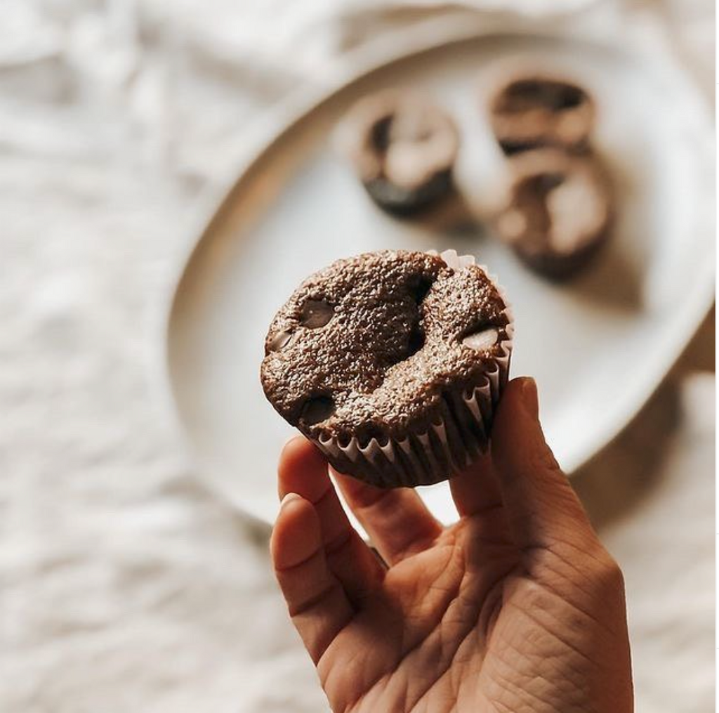 A Hand Holding a Muffin Made From Tejari Organic Cacao Superfood in the fore ground and in the back ground using a Blurry Filter the Remain Cacao Muffins on a Plate